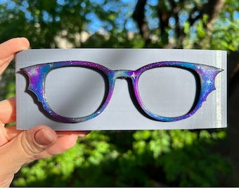 May include: A pair of galaxy-themed glasses with a purple, blue, and white design. The glasses have a cat-eye shape and are made of plastic.