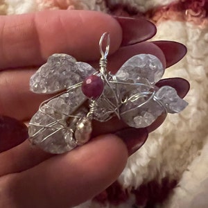 May include: A handmade pendant featuring a wire-wrapped design. The pendant showcases clear and light purple crystals, a small red gemstone, and silver-colored wire. The crystals are arranged in a wing-like shape, creating a unique and artistic piece of jewelry.