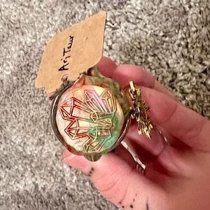May include: A decorative glass pendant with a crystal design in shades of red, green, and gold. The pendant is attached to a gold-colored metal clasp. A small tag with the word "Ari Taur" is attached to the pendant.