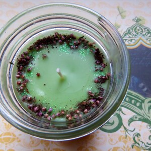 May include: A green candle in a clear glass jar. The candle is decorated with a ring of brown and purple dried flowers and green glitter. A white wick is centered in the candle. The candle sits on a patterned surface.