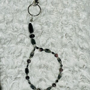 May include: A beaded keyring with a silver clasp and ring. The beads are a mix of black, grey, and pink, in various shapes and sizes. The keyring is designed to hold keys or other small items.