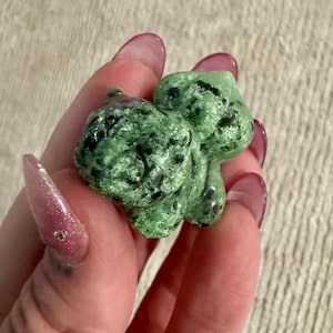 May include: A green stone carving of a small animal, possibly a rabbit or a bear. The stone is a mottled green with black inclusions. The carving is held in a hand with long, pink fingernails.