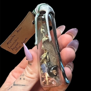 May include: A small glass vial containing dried flowers, crystals, and other natural elements. The vial has a decorative top with dripped paint. A small tag is attached to the vial with handwritten text. The vial is held against a black background.