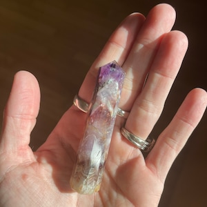 May include: A natural amethyst crystal point, featuring a gradient of purple, white, and brown hues. The crystal is held in a hand, showcasing its polished, elongated form. The crystal's tip is a deep purple, transitioning to lighter shades.