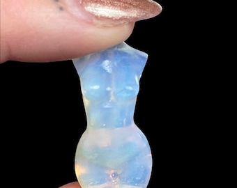 May include: A small, translucent, opalescent figurine of a female torso. The sculpture is held between two fingers against a black background. The figurine has a smooth, polished surface and a subtle blue and white color gradient.