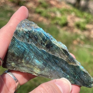 May include: A triangular piece of labradorite stone with iridescent blue and green hues. The stone has a rough, natural texture and is held in a hand. The background is blurred, showing green grass and trees.