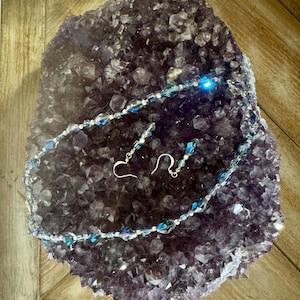 Könnte beinhalten: Ein Halsketten- und Ohrring-Set mit blauen und klaren Kristallperlen, präsentiert auf einer großen, dunkelvioletten Amethyst-Geode. Der Schmuck hat silberfarbene Metallbeschläge.