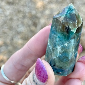 May include: A crystal point with blue, green, and brown hues. The polished crystal has a faceted shape. It is held in a hand with purple nail polish.