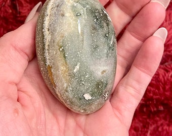 May include: A smooth, oval-shaped stone, displaying a blend of green, white, and tan hues. The polished surface reflects light, held in a hand. The stone's natural colours and patterns create an earthy aesthetic.