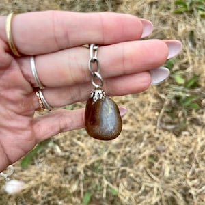 May include: A brown and white agate pendant with a silver chain. The teardrop-shaped stone has a polished surface and is suspended from a silver chain. The pendant is held in a hand.