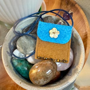May include: A small, handmade felt pouch with a blue flap and a flower-shaped button. The pouch is brown with a beaded trim and a blue cord. It rests on a bed of polished stones in a ceramic bowl.