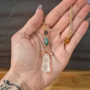 May include: A gold-coloured chain pendulum with a clear quartz crystal point and various gemstone beads. The pendulum features a clear quartz crystal point, a teal-coloured bead, and a gold-coloured chain.
