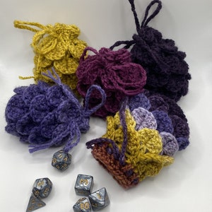 May include: A collection of handmade crochet dice bags in various colors, including gold, purple, and burgundy. Each bag has a drawstring closure and a textured, scale-like pattern. Several polyhedral dice are scattered nearby.