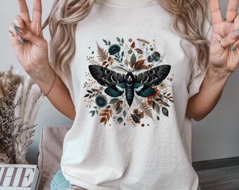 May include: A white t-shirt with a graphic print of a moth surrounded by flowers and leaves. The moth is blue, green, and black. The flowers are in shades of brown, orange, and blue.
