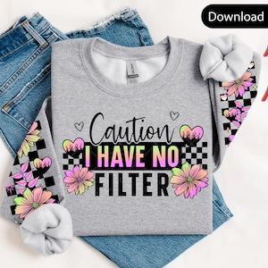 May include: A gray sweatshirt with the text "Caution I Have No Filter" in black and neon colors. The sleeves feature a checkered pattern with flowers and hearts in pink, yellow, and black. The sweatshirt is folded on top of blue jeans.