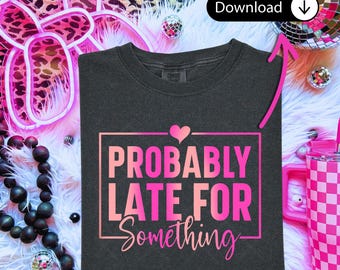 May include: Dark gray t-shirt with the text "PROBABLY LATE FOR Something" in pink and a pink heart. The shirt is folded on a fluffy white surface, with disco balls and a pink checkered tumbler in the background.