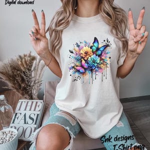 Puede incluir: Una camiseta blanca con un diseño de acuarela colorido de mariposas y flores. El diseño tiene un efecto de goteo e incluye tonos de azul, rosa, amarillo y verde.