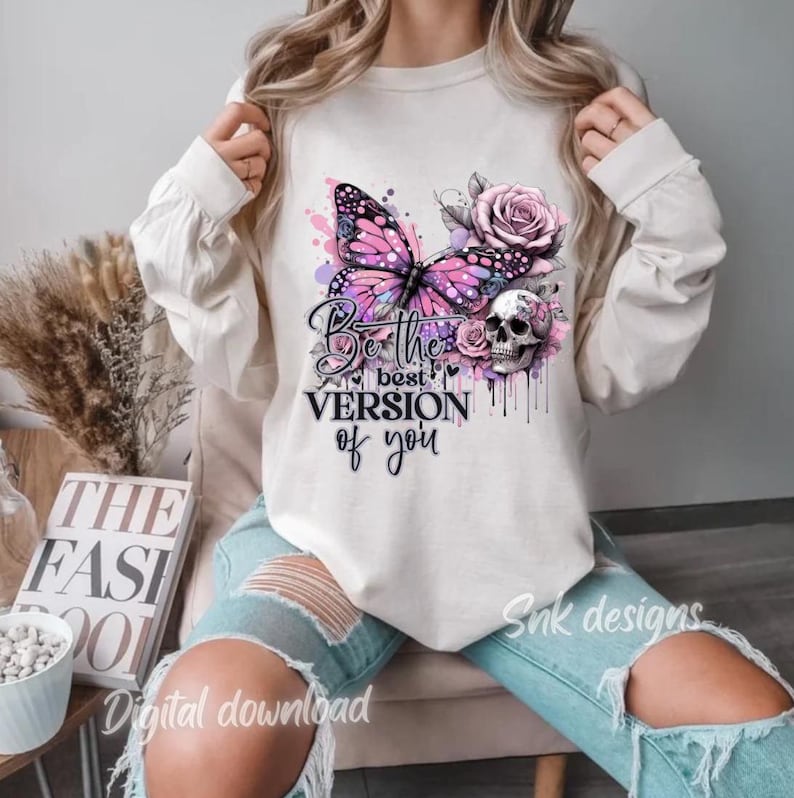 May include: A white sweatshirt featuring a pink butterfly, skull, and flowers. The text "Be the best version of you" is printed on the front of the sweatshirt.