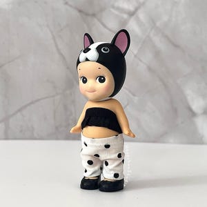 K&ouml;nnte beinhalten: Eine kleine Figur mit einer schwarz-wei&szlig;en Franz&ouml;sische-Bulldogge-M&uuml;tze mit rosa Innenohren. Die Figur tr&auml;gt ein schwarzes tr&auml;gerloses Oberteil mit ger&uuml;schtem Rand und eine wei&szlig;e Hose mit schwarzem Punktemuster. An den F&uuml;&szlig;en tr&auml;gt sie schwarze Schuhe.