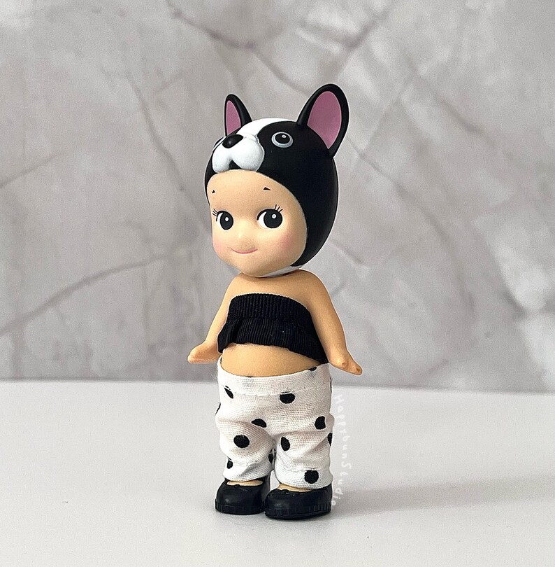 K&ouml;nnte beinhalten: Eine kleine Figur mit einer schwarz-wei&szlig;en Franz&ouml;sische-Bulldogge-M&uuml;tze mit rosa Innenohren. Die Figur tr&auml;gt ein schwarzes tr&auml;gerloses Oberteil mit ger&uuml;schtem Rand und eine wei&szlig;e Hose mit schwarzem Punktemuster. An den F&uuml;&szlig;en tr&auml;gt sie schwarze Schuhe.