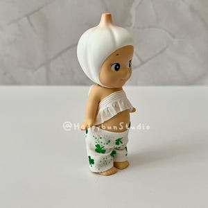 May include: A small figurine of a baby-like character with a garlic bulb head. The figurine wears a white ruffled top and green pants with shamrock patterns. The figurine is standing on a white surface.