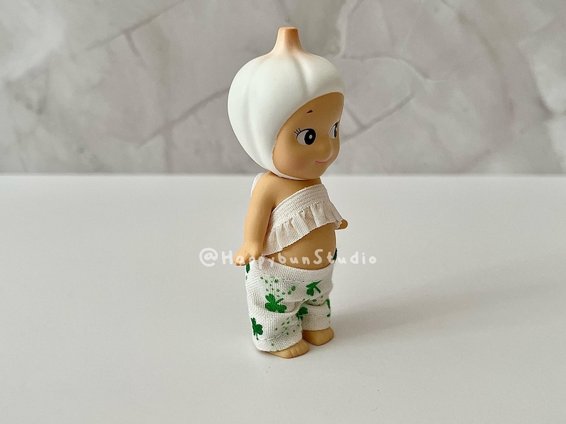 May include: A small figurine of a baby-like character with a garlic bulb head. The figurine wears a white ruffled top and green pants with shamrock patterns. The figurine is standing on a white surface.