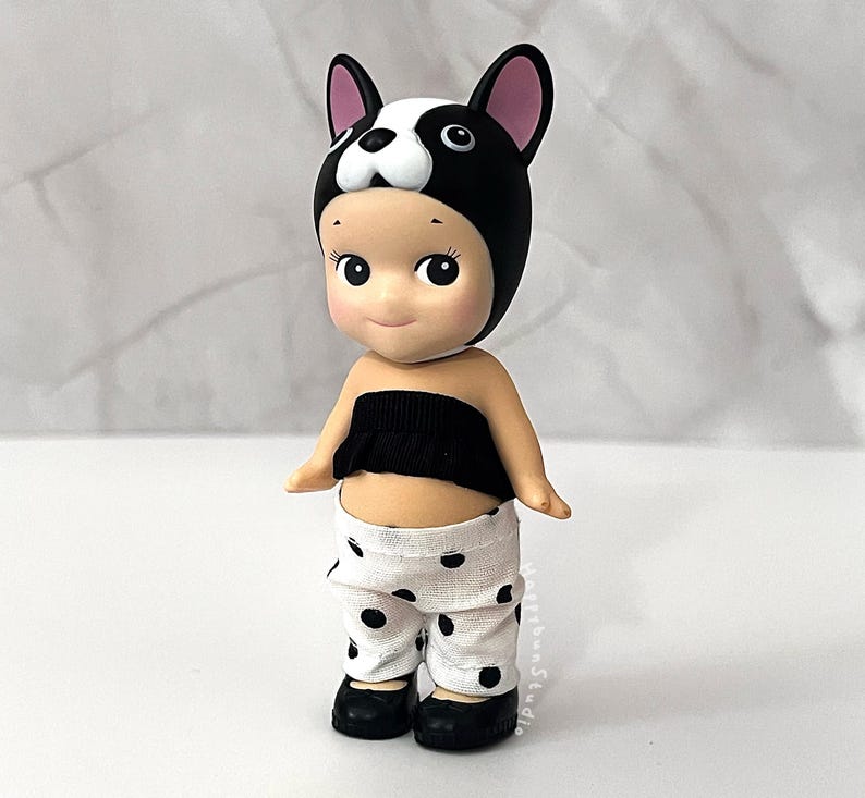 K&ouml;nnte beinhalten: Eine kleine Sammelfigur mit einer schwarz-wei&szlig;en Franz&ouml;sische Bulldogge-Kopfbedeckung, einem schwarzen tr&auml;gerlosen Oberteil und einer wei&szlig;en Hose mit schwarzen Punkten. Die Figur hat helle Haut, gro&szlig;e dunkle Augen und tr&auml;gt kleine schwarze Schuhe.