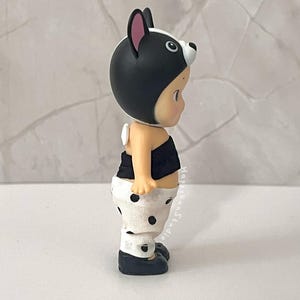 K&ouml;nnte beinhalten: Eine kleine Figur mit einer schwarz-wei&szlig;en Kapuze im Tierdesign, einem schwarzen tr&auml;gerlosen Oberteil und einer wei&szlig;en Hose mit schwarzen Punkten. Die Figur hat einen hellen Hautton und tr&auml;gt dunkle Schuhe. Der Text HappyBunStudio ist vertikal gedruckt.