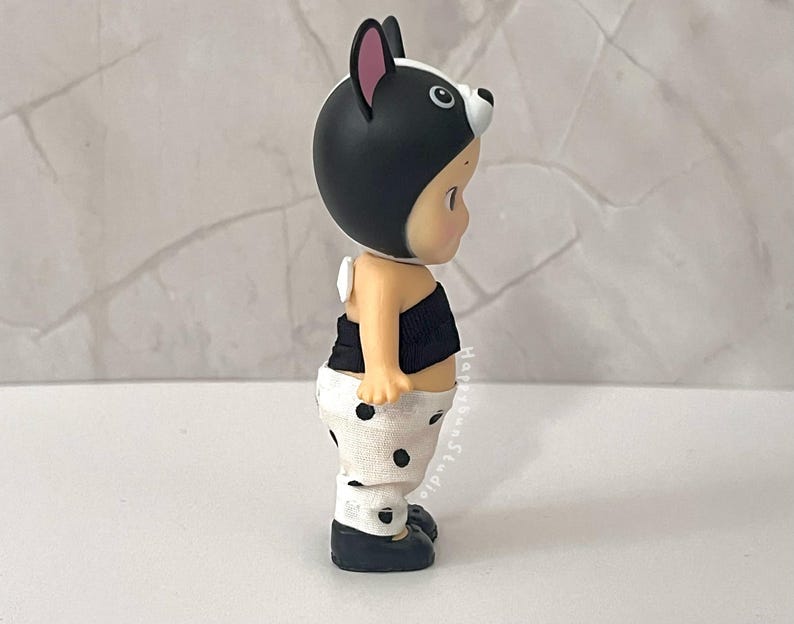 K&ouml;nnte beinhalten: Eine kleine Figur mit einer schwarz-wei&szlig;en Kapuze im Tierdesign, einem schwarzen tr&auml;gerlosen Oberteil und einer wei&szlig;en Hose mit schwarzen Punkten. Die Figur hat einen hellen Hautton und tr&auml;gt dunkle Schuhe. Der Text HappyBunStudio ist vertikal gedruckt.
