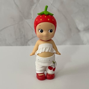 May include: A small figurine of a doll wearing a strawberry hat and red shoes. The doll has a white ruffled top and white pants with cherry designs. The strawberry hat is red with yellow dots and a green stem.