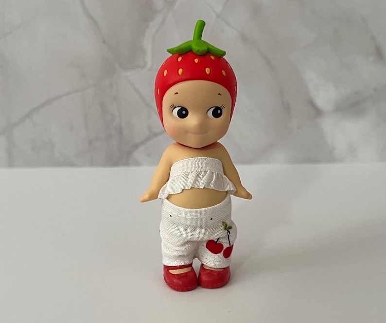 May include: A small figurine of a doll wearing a strawberry hat and red shoes. The doll has a white ruffled top and white pants with cherry designs. The strawberry hat is red with yellow dots and a green stem.