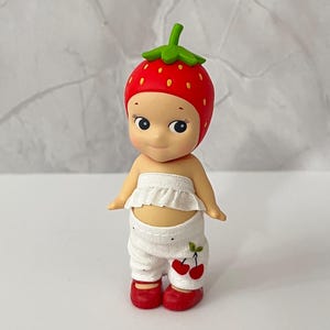 May include: A small figurine of a doll wearing a red strawberry hat with green leaves, a white ruffled top, and white pants with a cherry design. The doll has red shoes and a smiling expression.