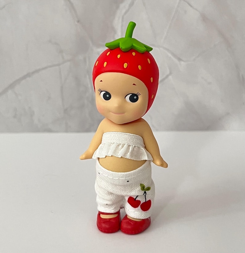 May include: A small figurine of a doll wearing a red strawberry hat with green leaves, a white ruffled top, and white pants with a cherry design. The doll has red shoes and a smiling expression.