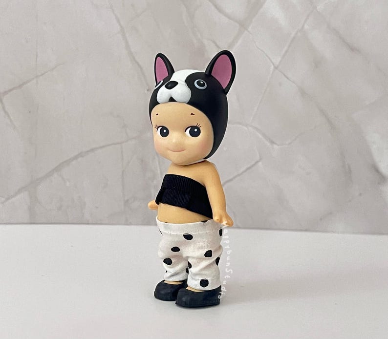 K&ouml;nnte beinhalten: Eine kleine Figur mit einer schwarz-wei&szlig;en Franz&ouml;sische Bulldogge-Kopfbedeckung, einem schwarzen tr&auml;gerlosen Oberteil und einer wei&szlig;en Hose mit schwarzen Punkten. Die Figur hat einen hellen Hautton, rosige Wangen und schwarze Schuhe.