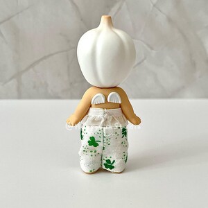 May include: A whimsical figurine with a white bulbous head and small, cream-colored wings. The figurine has tan arms and is wearing white pants with green shamrock patterns. The figure is a unique decorative item.