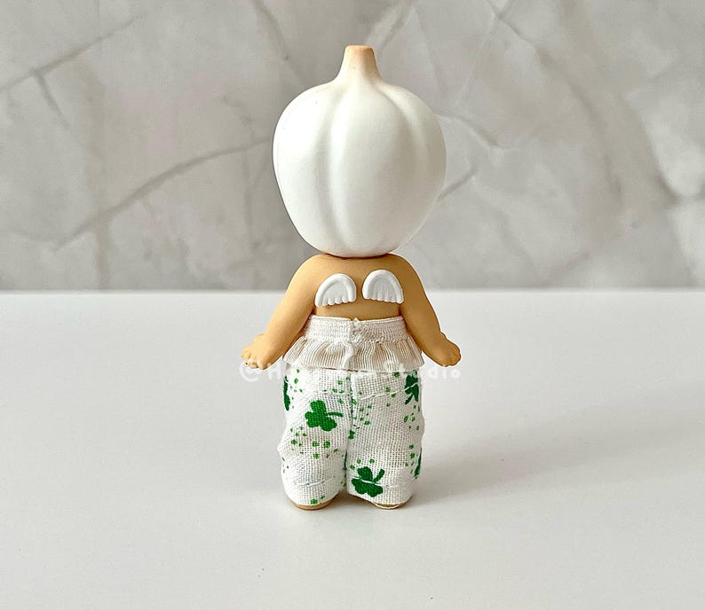 May include: A whimsical figurine with a white bulbous head and small, cream-colored wings. The figurine has tan arms and is wearing white pants with green shamrock patterns. The figure is a unique decorative item.