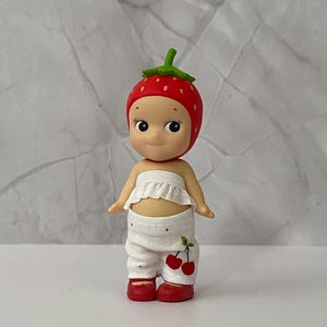 May include: A small figurine of a doll wearing a red strawberry hat with white polka dots. The doll has a white ruffled top and white pants with a cherry design. The doll is wearing red shoes.