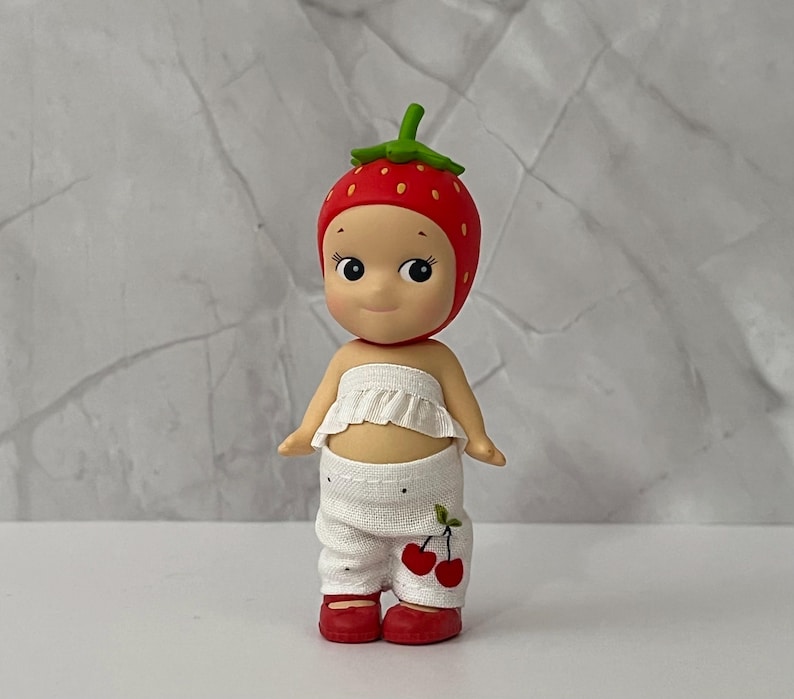May include: A small figurine of a doll wearing a red strawberry hat with white polka dots. The doll has a white ruffled top and white pants with a cherry design. The doll is wearing red shoes.