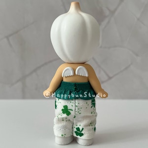 May include: A whimsical figurine with a white bulbous head and tan body. It wears a green top and white pants patterned with green shamrocks. Small white wings are attached to its back. The figurine is standing.