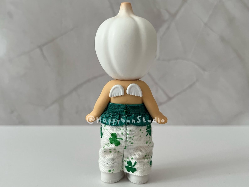 May include: A whimsical figurine with a white bulbous head and tan body. It wears a green top and white pants patterned with green shamrocks. Small white wings are attached to its back. The figurine is standing.