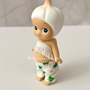 May include: A whimsical figurine of a baby-like character wearing a white garlic bulb hat and a ruffled top. The figure has a smiling face and is wearing white pants with green shamrock patterns. The figurine is a collectible item.