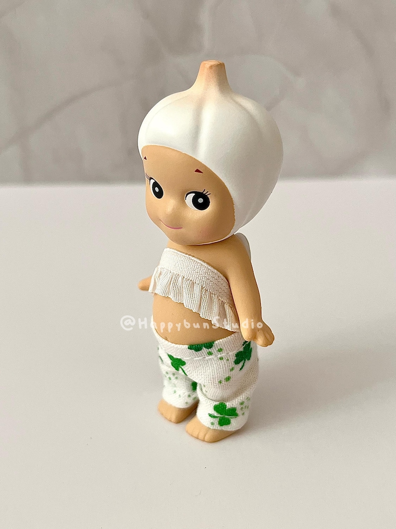 May include: A whimsical figurine of a baby-like character wearing a white garlic bulb hat and a ruffled top. The figure has a smiling face and is wearing white pants with green shamrock patterns. The figurine is a collectible item.
