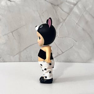 K&ouml;nnte beinhalten: Eine kleine Figur mit einer schwarzen Kapuze mit rosa gef&uuml;tterten Ohren, einem schwarzen tr&auml;gerlosen Oberteil und einer wei&szlig;en Hose mit schwarzem Punktemuster. Die Figur hat einen hellen Hautton und ist im Seitenprofil vor Marmor zu sehen.