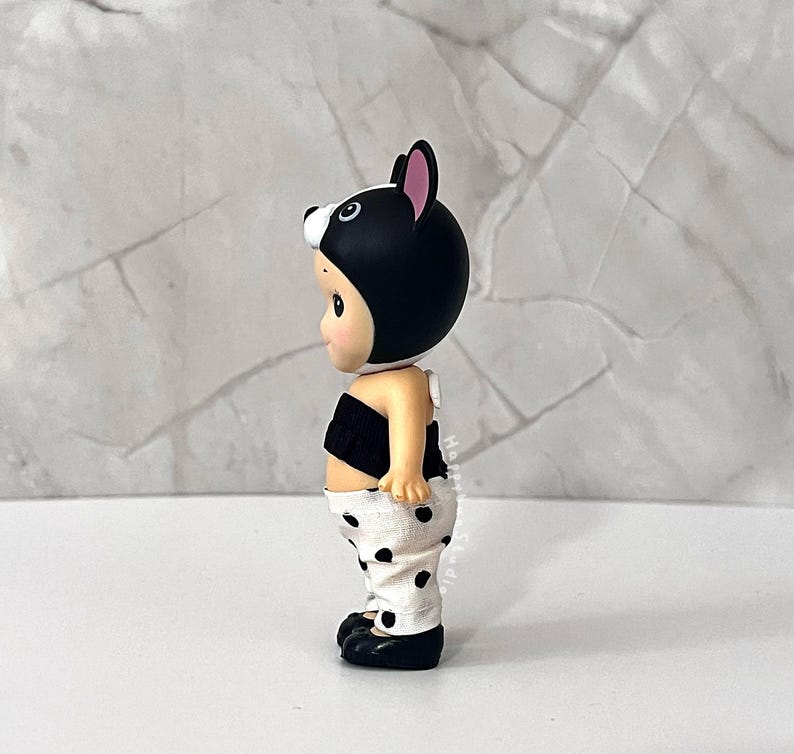 K&ouml;nnte beinhalten: Eine kleine Figur mit einer schwarzen Kapuze mit rosa gef&uuml;tterten Ohren, einem schwarzen tr&auml;gerlosen Oberteil und einer wei&szlig;en Hose mit schwarzem Punktemuster. Die Figur hat einen hellen Hautton und ist im Seitenprofil vor Marmor zu sehen.