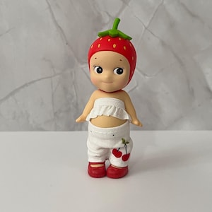 May include: A small figurine of a baby wearing a strawberry hat, white ruffled top, and white pants with cherry accents. The figurine has red shoes and a sweet expression.