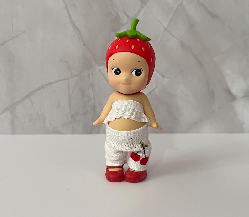 May include: A small figurine of a baby wearing a strawberry hat, white ruffled top, and white pants with cherry accents. The figurine has red shoes and a sweet expression.