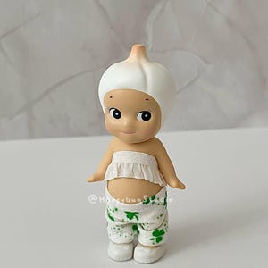 May include: A small, whimsical figurine of a character wearing a white garlic bulb hat and a ruffled white top. The character is also wearing white pants with green shamrock patterns. The figurine has a smiling face and small arms.