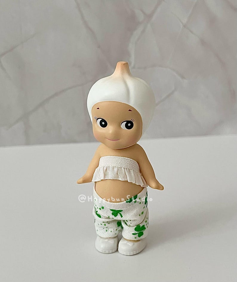 May include: A small, whimsical figurine of a character wearing a white garlic bulb hat and a ruffled white top. The character is also wearing white pants with green shamrock patterns. The figurine has a smiling face and small arms.