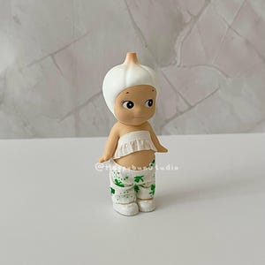 May include: A small figurine of a character dressed as a garlic bulb. The figure has a white garlic bulb head, a ruffled white top, and green and white patterned pants. The figure is standing on a white surface.