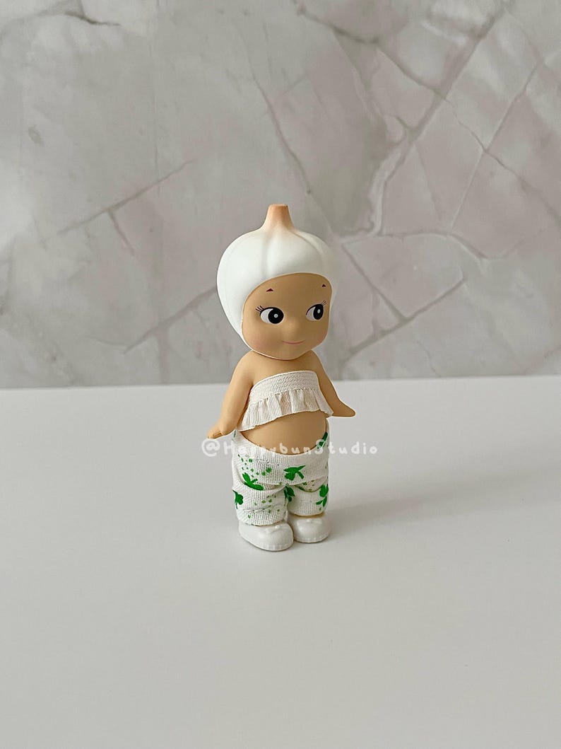 May include: A small figurine of a character dressed as a garlic bulb. The figure has a white garlic bulb head, a ruffled white top, and green and white patterned pants. The figure is standing on a white surface.